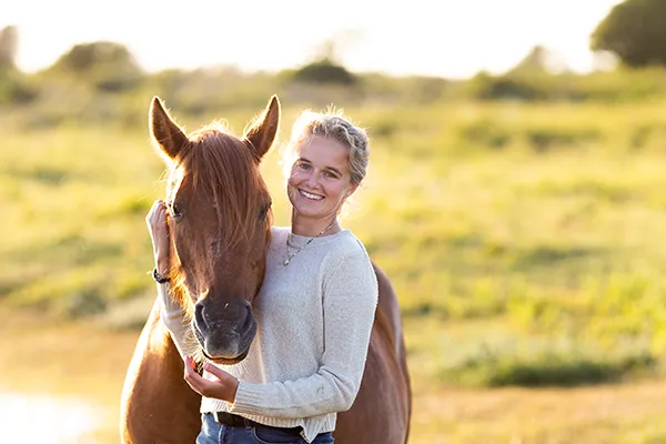 Une femme souriante en pull beige enlace un cheval alezan dans un cadre champêtre baigné de lumière douce, symbolisant une connexion sincère et harmonieuse.