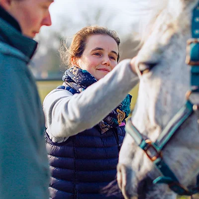 Découvrir le shiatsu. Une praticienne en shiatsu sourit et applique un toucher doux sur la tête d'un cheval gris portant un licol bleu. Une autre personne observe la scène, créant un moment de partage et d'apprentissage.